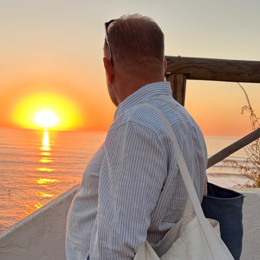 A man in a striped linen shirt watching the sun set over the sea from a whitewashed terrace