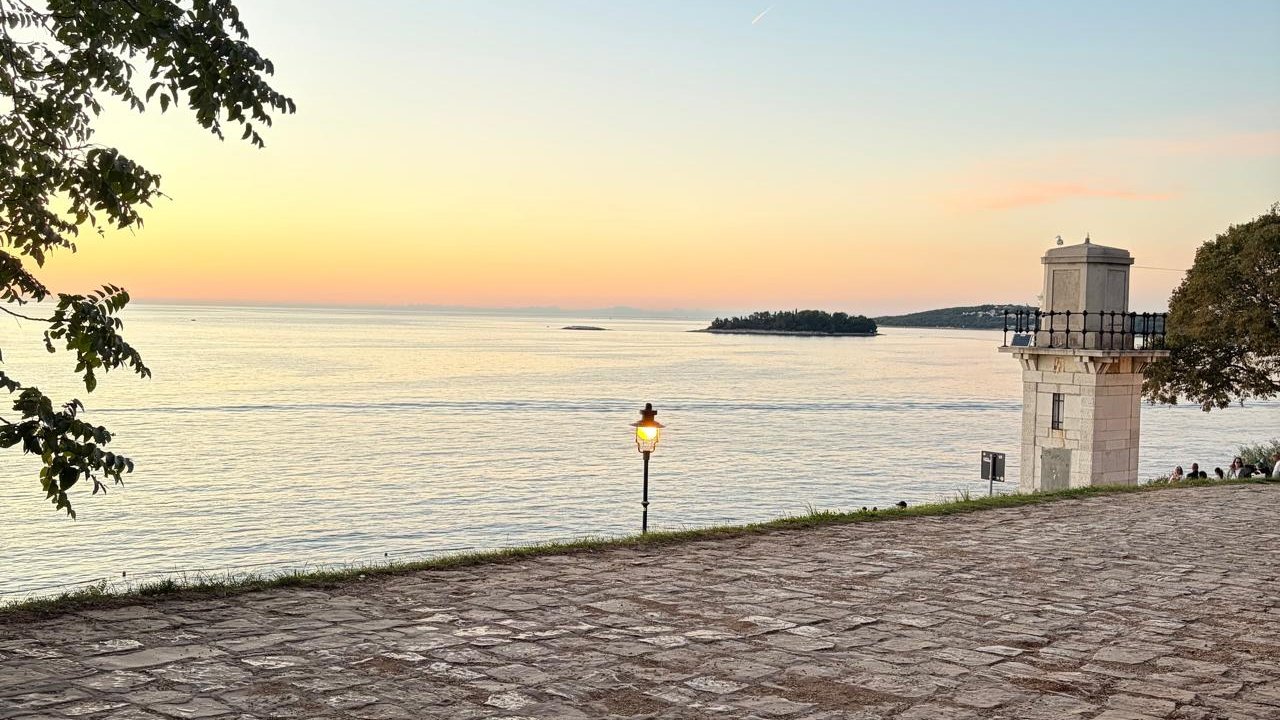 A cobbled promenade at sunset with a small lighthouse and a glowing lamp, looking out over the Adriatic from Rovinj